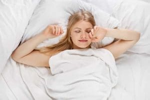 A woman lying in bed with a sheet covering her, her arms outstretched while her hands are pointed in towards her face.